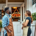 A black man and white woman both wear aprons and are outside of their small business and t