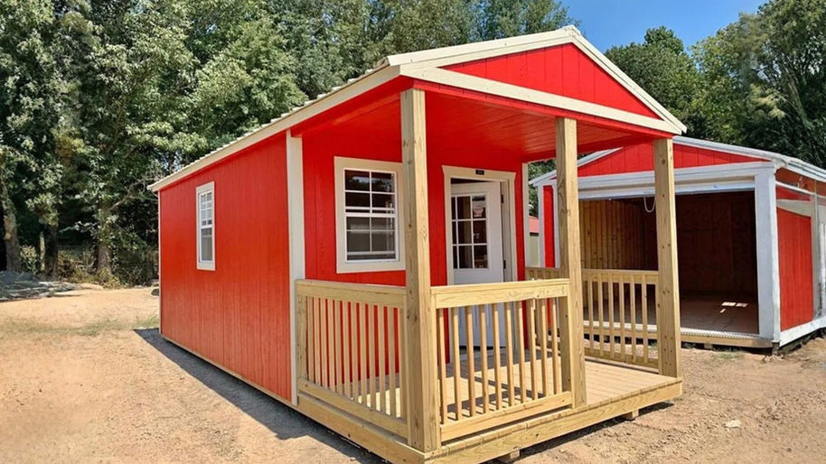 A bright red, portable cabin with white trim is set outdoors on a dirt or gravel lot, surrounded by green trees in the background. The cabin is 12ft×28ft and features an end porch with natural wood railings, flooring, and support posts. There is a white-framed window on the left side of the porch and a white exterior door leading into the cabin. The roof overhangs the porch slightly. Another similar, but larger, red and white structure with an open front is visible to the right.