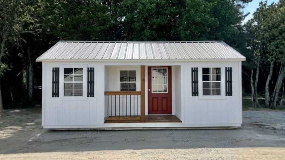 A white, rectangular portable cabin measuring $12 \text{ft} \times 24 \text{ft}$ is centered in the frame against a backdrop of dense green trees and a dirt/gravel foreground. The cabin has a corrugated silver metal roof and features a small center porch. The porch has a dark, reddish-brown wooden floor, a black metal railing on the left side, and is framed by two white windows with black shutters on either side. The main entrance is a bright red door with a partial glass insert, flanked by a smaller window pane to the left.