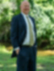 A front-view image of Guy Wade smiling in a suit with a gold tie in front of green trees