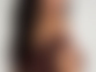 Eine junge Frau mit langen, dunklen Wellen und Pony posiert in einem eng anliegenden, schokobraunen Off-Shoulder-Kleid. Sie blickt über ihre Schulter in die Kamera. Das Kleid hat einen leicht ausgestellten Rockteil. Sie trägt mehrere Armreifen an beiden Handgelenken vor einem hellen, minimalistischen Hintergrund.