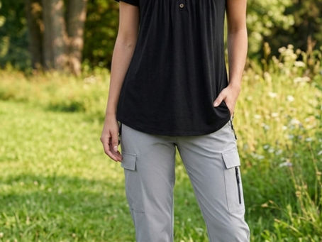 Eine lächelnde Frau mit brünetten, welligen Haaren steht auf einer grünen Wiese vor einem Hintergrund aus Bäumen. Sie trägt ein schwarzes, locker fallendes Kurzarm-Shirt mit einer kleinen Knopfleiste am Ausschnitt, eine hellgraue Cargo-Hose mit Reißverschlusstaschen und weißen Sneakern. Die Lichtstimmung ist sonnig und natürlich.