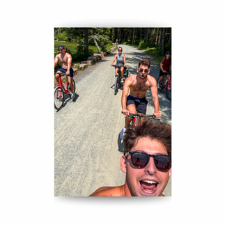 Selfie of four cyclists on a sunny forest gravel road, including shirtless men and a woman, riding and smiling.