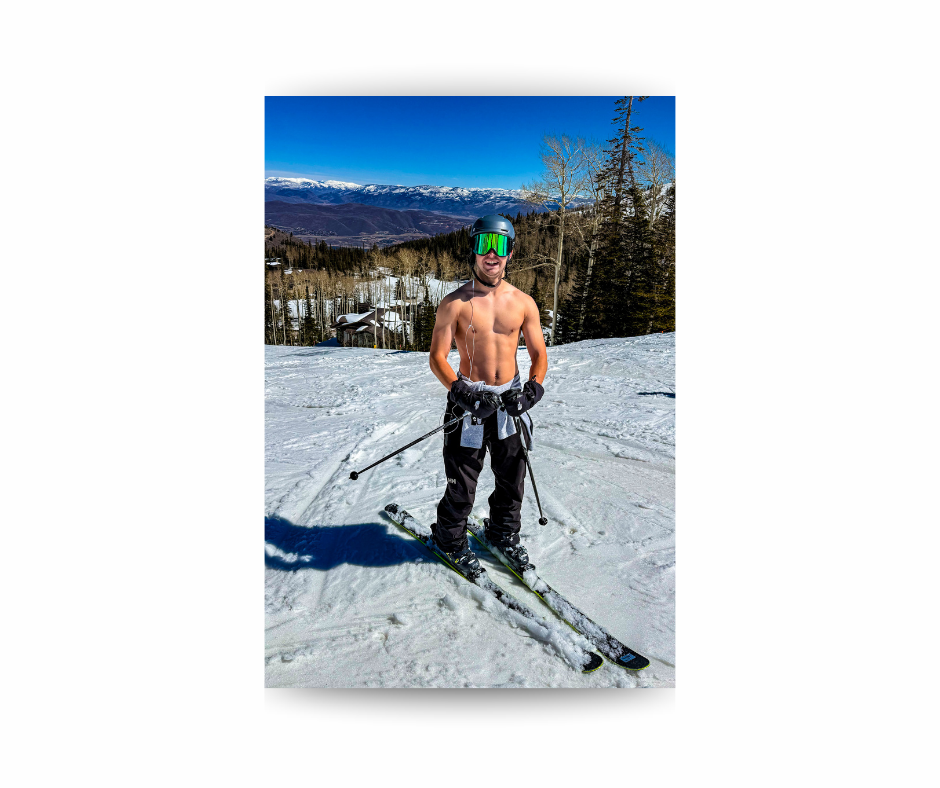 Man skiing shirtless on a snowy slope, wearing ski pants and goggles. Forest and mountains in the background under a clear blue sky.