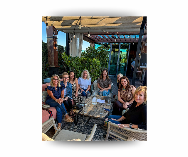 Group of midlife women gathered outdoors around a table, smiling and enjoying conversation during a wellness-focused meetup that celebrates friendship and community connection.