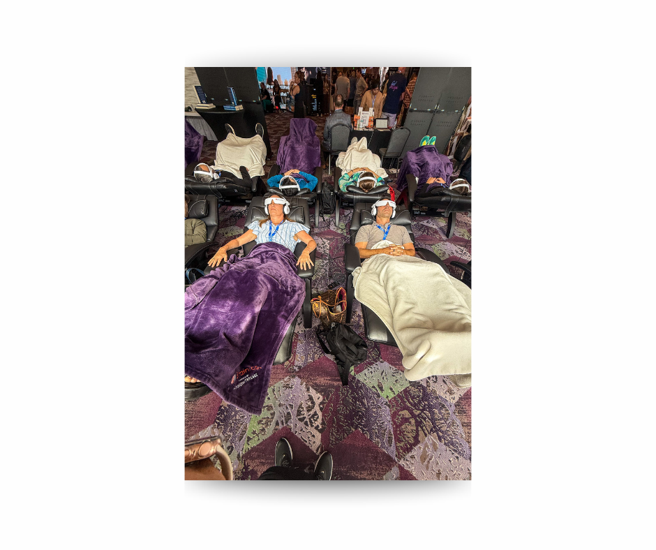 People recline in massage chairs under purple and cream blankets at a busy expo, with booths and crowds in the background.