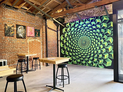 The photo depicts an interior space of the brewery with an industrial aesthetic featuring a brick wall on the left adorned with four eclectic paintings. In the foreground, there are wooden tables with black stools. Dominating the right side is a striking, large, green geometric mural creating an optical illusion of depth. Exposed wooden beams and ductwork are visible overhead, and glass doors with natural light are on the right