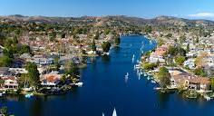 Aerial view of a calm river flanked by houses and trees. Small boats sail on the blue water. Rolling hills under a clear blue sky in the background.