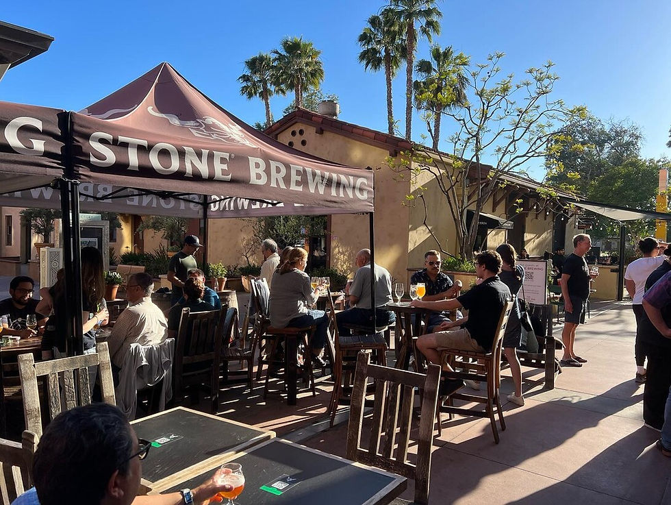 People enjoy drinks under a "Stone Brewing" tent outside on a sunny day with palm trees and warm-colored buildings in the background.