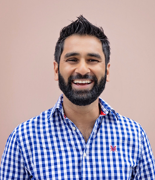 Amit stands against a soft pink backdrop, smiling warmly with a relaxed, confident posture. He wears a blue and white checkered shirt with a small red emblem on the chest, his dark hair styled upward and a full beard framing his face.