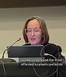 Video frame showing Barbra “Barbie” Weber seated at a long table indoors and speaking into a microphone with a laptop in front of her. She is wearing glasses and a black suit jacket. Behind her is a plain gray wall. The caption from this part of her INC-4 speech showing up in the frame reads, "waste pickers and the most affected by plastic pollution"