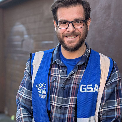 Portrait photo of Carlos, a Ground Score worker with short brown hair, a short brown beard, black-framed eyeglasses, a blue checked flannel shirt, and a blue Ground Score vest, smiling and looking at the camera.