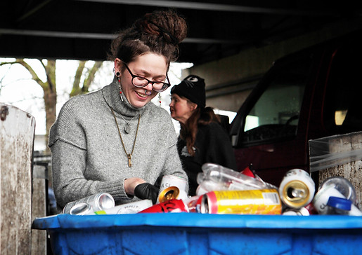 Photo of a Ground Score worker smiling and sorting empty bottles and cans for recycling at The People's Depot in Portland, Oregon. She is wearing a gray sweater, long earrings, two pendants, black-framed glasses, and black work gloves. Her brown hair is tied in a bun on the top of her head. In the background is another worker, multiple tall rectangular boxes, a dark-colored vehicle, and the underside of the bridge the depot is located under. Beyond the bridge is a tree.