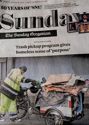 Photo of front page of The Sunday Oregonian showing that the main story is  about Ground Score. The title is 'Trash pickup program gives homeless sense of "purpose."' Underneath the headline is a large photo of a Ground Score worker wearing a neon yellow reflective jacket and pants standing next to an adult trike with a rectangular bin mounted in the front for hauling waste. The bin is lined with a gray tarp and filled with pieces of cardboard strapped together. The worker is walking on a paved area in front of a gray building exterior. At the very top of the newspaper's front page, it also says "50 years of SNL" and includes three images inside TV graphics from the show.