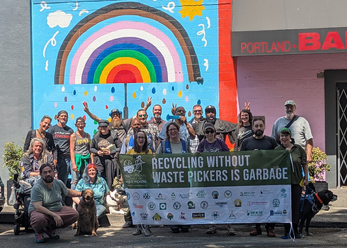 Group photo of about twenty Ground Score workers and three dogs posing outside on a sunny day. The people on the right side are holding a large banner in front of them that says "Recycling without waste pickers is garbage" in large letters in white on the top. The banner has a dark green top behind that text and the logo of the International Alliance of Waste Pickers next to the text. The bottom part of the banner is white with the logos of various waste picker organizations from around the world.  Behind the group of workers is an outdoor wall with a mural of a progress pride rainbow shaped like an umbrella with colorful raindrops coming down below it. The mural also features sun and cloud elements above the rainbow umbrella and a light blue background behind it. To the right of that area of the mural is an area of red-orange next to an area of purplish pink with a black awning over a door; the awning says in red letters “Portland BA” with the part after that cut off.