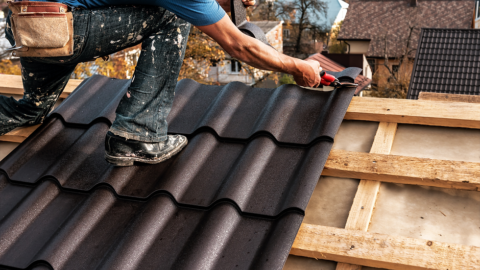 Person in blue shirt and jeans installing dark roof tiles with red tool. Wooden beams visible beneath, autumn trees in background.