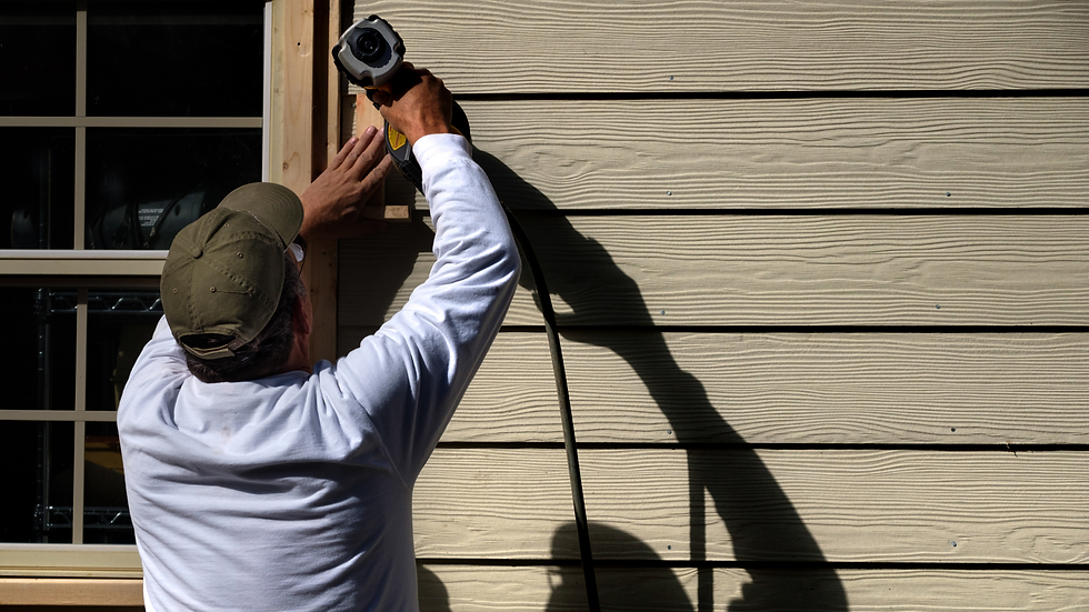 Man in a cap using a drill on wood siding, casting a shadow. Sunlit beige panels in the background. Daytime, focused work.