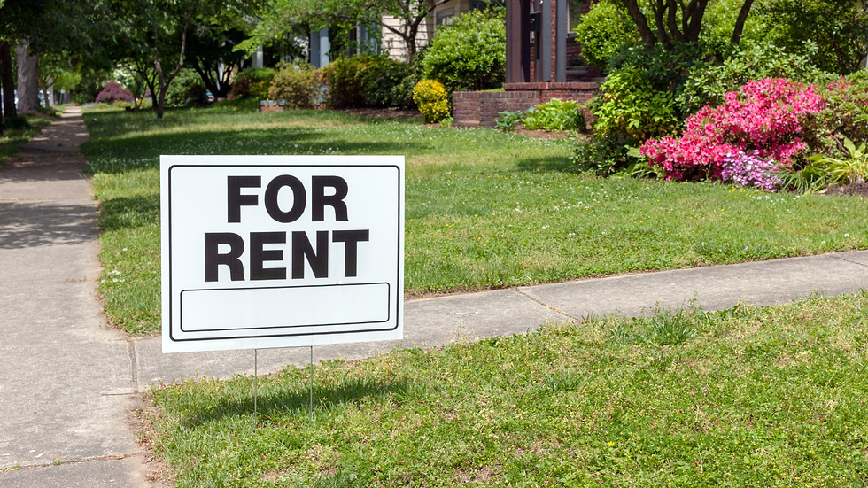 For rent sign on a grassy suburban lawn beside a sidewalk, with trees, brick houses, and pink flowers in the background.