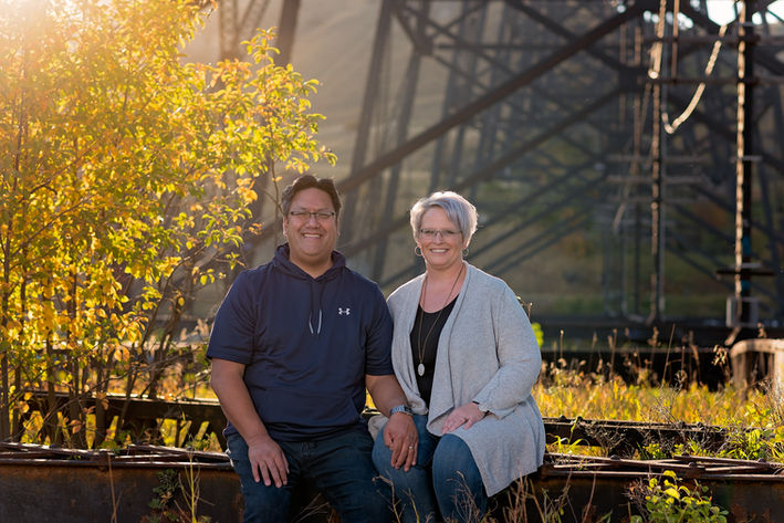 Couple sitting together outdoors near railway tracks and a steel bridge surrounded by golden fall foliage.