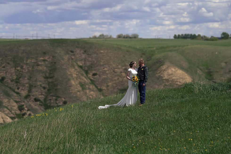 Bride and groom stand on a grassy hill, gazing at each other, the bride holding a sunflower bouquet under a cloudy sky.