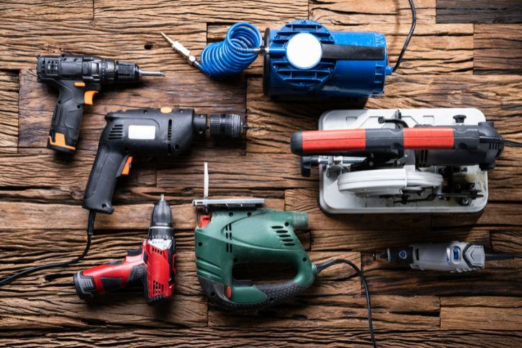 Various power tools, including drills and a saw, are arranged on a rustic wooden surface. Cables and a blue coil are visible.