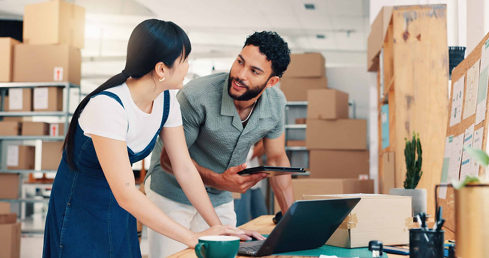 Two people collaborate in an office with boxes. One uses a laptop, the other holds a tablet. The setting is organized and friendly.
