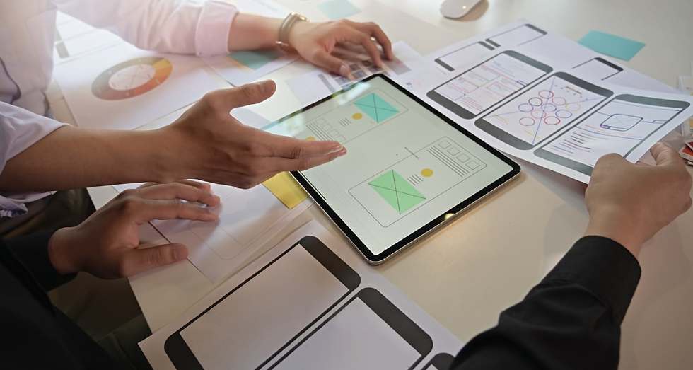 Hands discussing wireframe designs on a tablet and papers in an office setting. Bright, organized workspace with digital and paper elements.