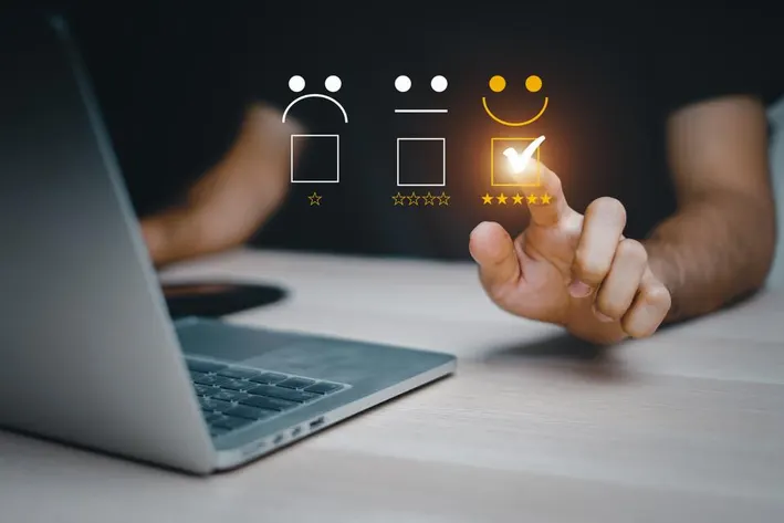 A person selects a happy face rating on a digital screen beside a laptop. The background is dark, emphasizing the glowing checkmark and stars.