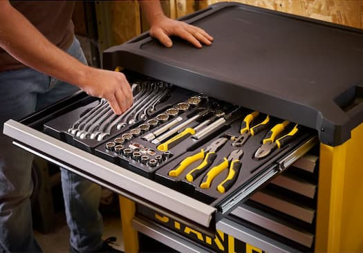 Person organizing hand tools in a black and yellow Stanley tool cabinet drawer, inside a wooden workshop. Tools include wrenches and pliers.