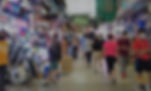 People wearing masks shop in a bustling indoor market. Bright stalls line the aisles with colorful lights and goods. Signs and boxes are visible.