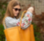 Woman with sunglasses adjusts striped floral sweater, holds large orange handbag. Brick wall and greenery in background; casual mood.