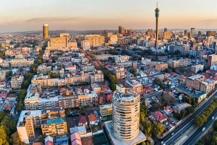 Aerial view of a bustling city at sunset, tall buildings and a prominent tower in the foreground, with roads lined by trees and houses.