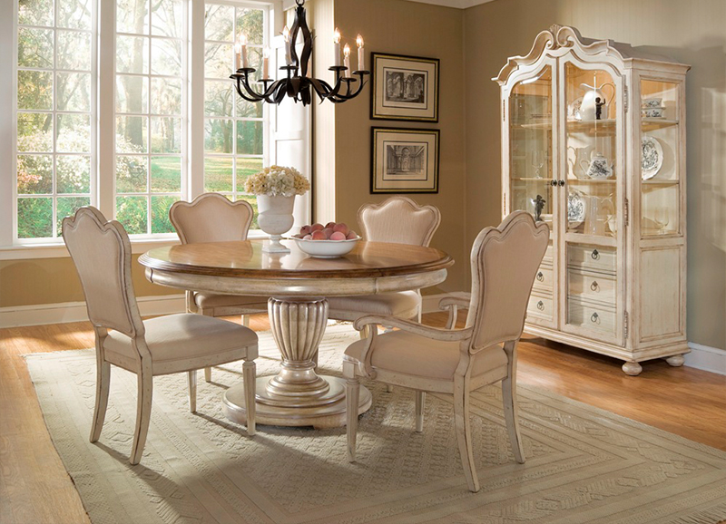 Elegant dining room with a round table, four beige chairs, a chandelier, and a china cabinet. Large windows reveal a sunny garden view.