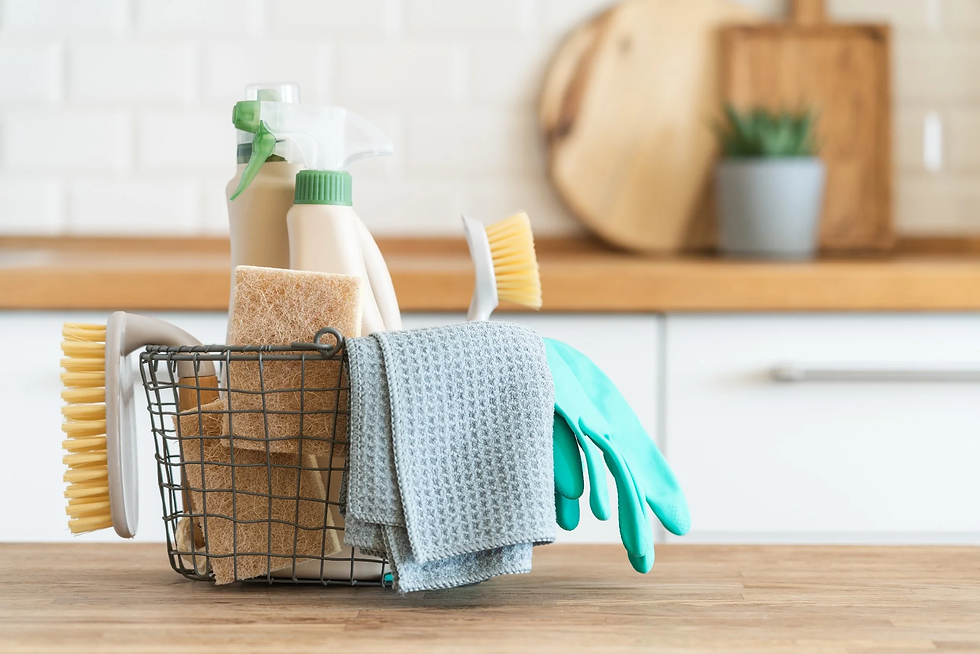 Cleaning supplies in a wire basket on a wooden countertop. Includes sponges, brushes, cloth, bottles, and teal gloves. White tiled background.