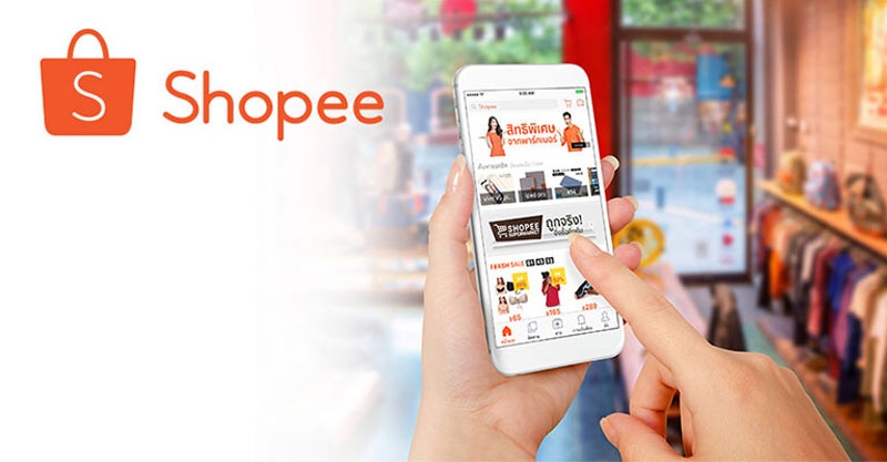 Hand holding a smartphone with the Shopee app open, showing sale items. Shopee logo on left. Blurred store background, warm lighting.