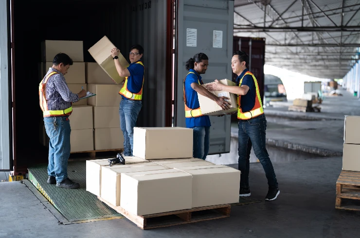 Warehouse workers in safety vests load boxes onto a pallet in a large storage area. The setting is industrial, with open metal roofing.