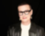 A photograph of a woman with short hair and chunky black eyeglasses. She is smiling and wearing a leather jacket.