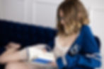 Woman in glasses reading a blue book on a navy sofa, wearing a blue jacket with an emblem and white lace attire. Intimate and personalized boudoir photoshoot in Minnesota.
