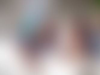 Two people with tattoos lie on a bed, one with rainbow hair uses a camera, the other holds a phone. A large mirror and plants are in the background.