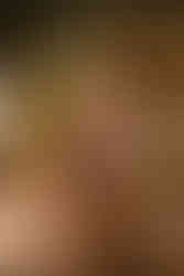 A woman stands leaning against a wooden table covered in candles. The background is a dark green wall filled with golden frame art and a mirror. 