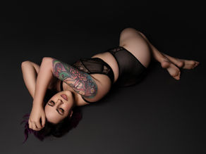 A woman lies on a dark floor, wearing black lingerie, showing a colorful arm tattoo. She appears relaxed. Dark background.