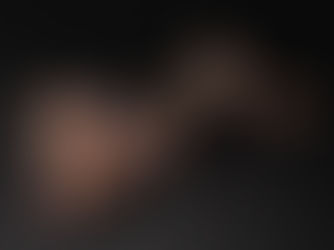 A woman lies on a dark floor, wearing black lingerie, showing a colorful arm tattoo. She appears relaxed. Dark background.