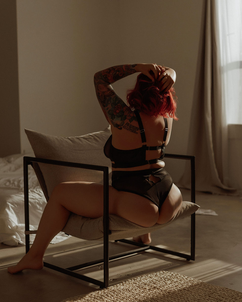 A person with red hair sits on a chair facing away, holding their head. Tattoos cover one arm. Sunlight filters through a window in a calm room.