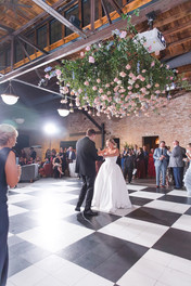 Bride and groom dancing under hanging flowers on black and white checkered dance floor.