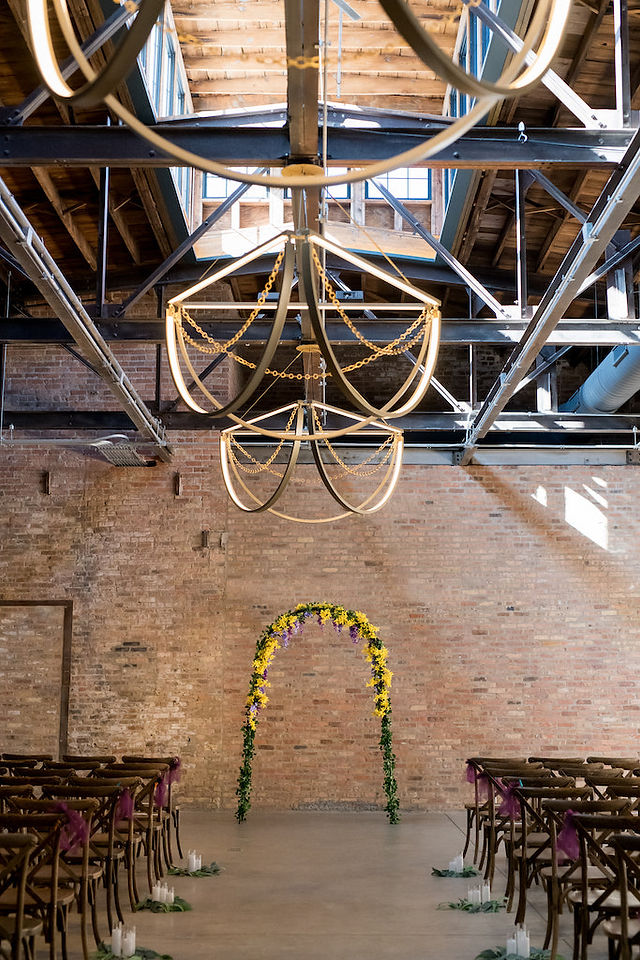 Indoor wedding ceremony setup with yellow and purple floral arch at industrial venue.