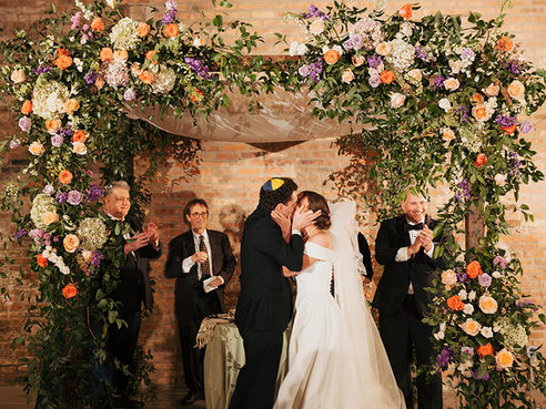 Bride and groom kiss beneath floral chuppah at industrial Chicago wedding venue.