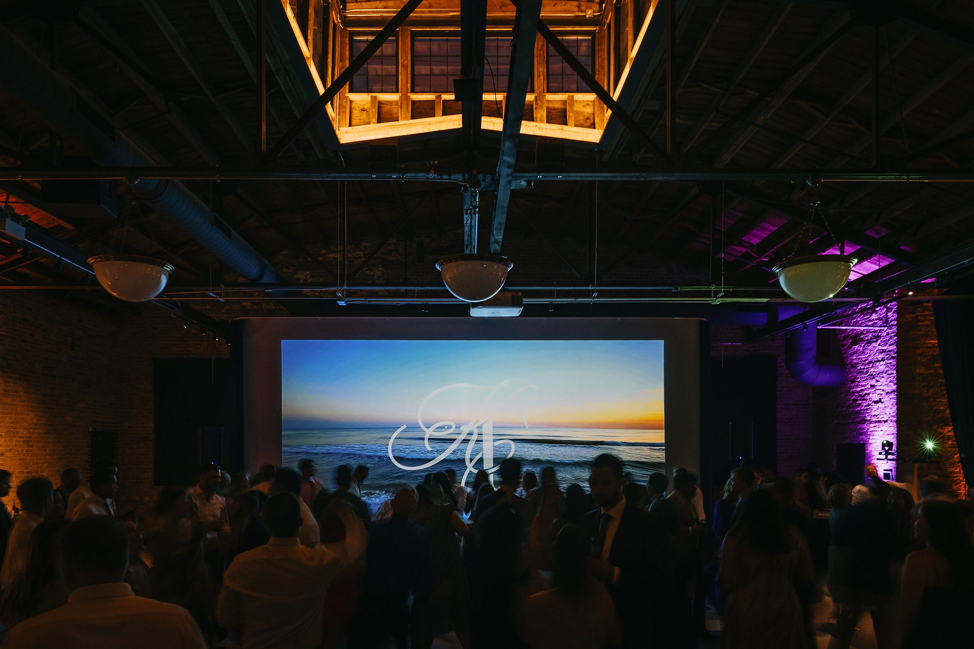 Guests dance in front of a custom monogram projection and ocean sunset visual at Wildman BT wedding reception.