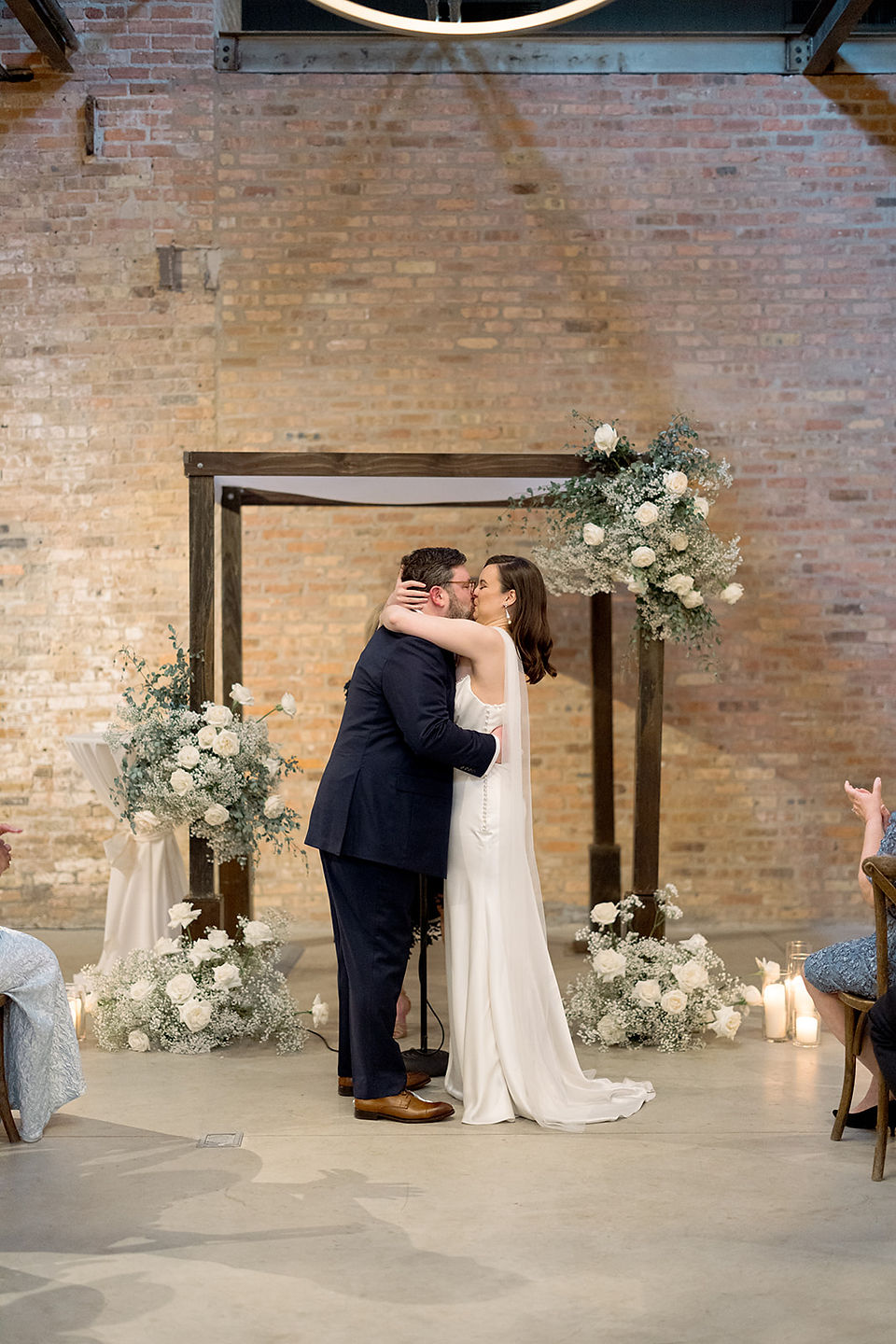 Bride and groom share kiss beneath wooden arch with white florals.