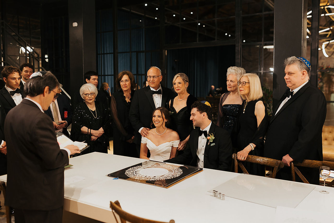 Jewish wedding signing ceremony with bride, groom, and family gathered indoors.