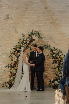 Bride and groom exchange vows beneath floral arch.